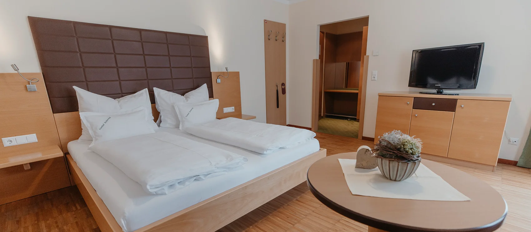 Blick in ein Zimmer - links befindet sich ein großes Doppelbett, rechts ein kleiner runder Tisch und an der Wand steht eine Kommode mit einem TV-Gerät.